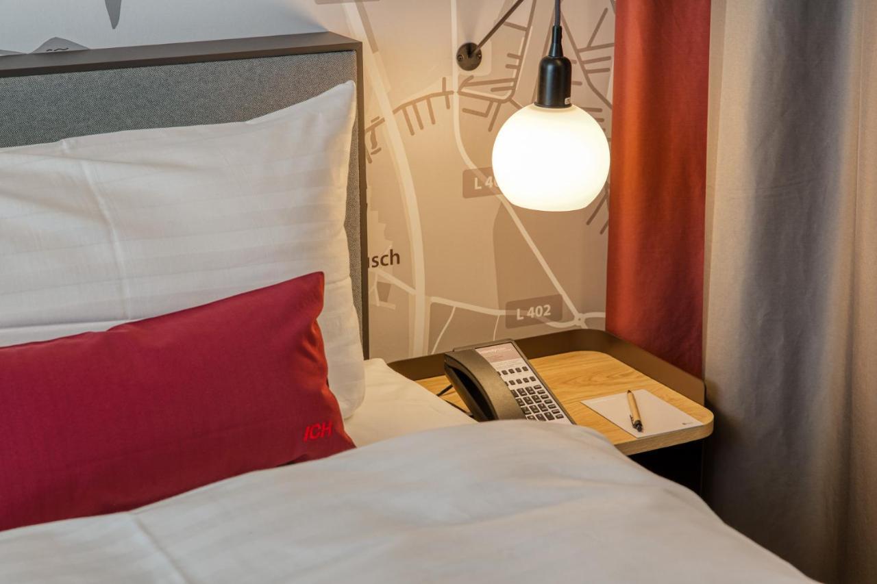 Intercityhotel Berlin Airport Ber Terminal 1 & 2 Schonefeld Bagian luar foto The photo shows a cozy bedroom setup. There's a neatly made bed with white bedding and a red decorative pillow. Beside the bed, a small bedside table holds a telephone and a notebook or notepad, along with a pen. A round pendant light hangs above the